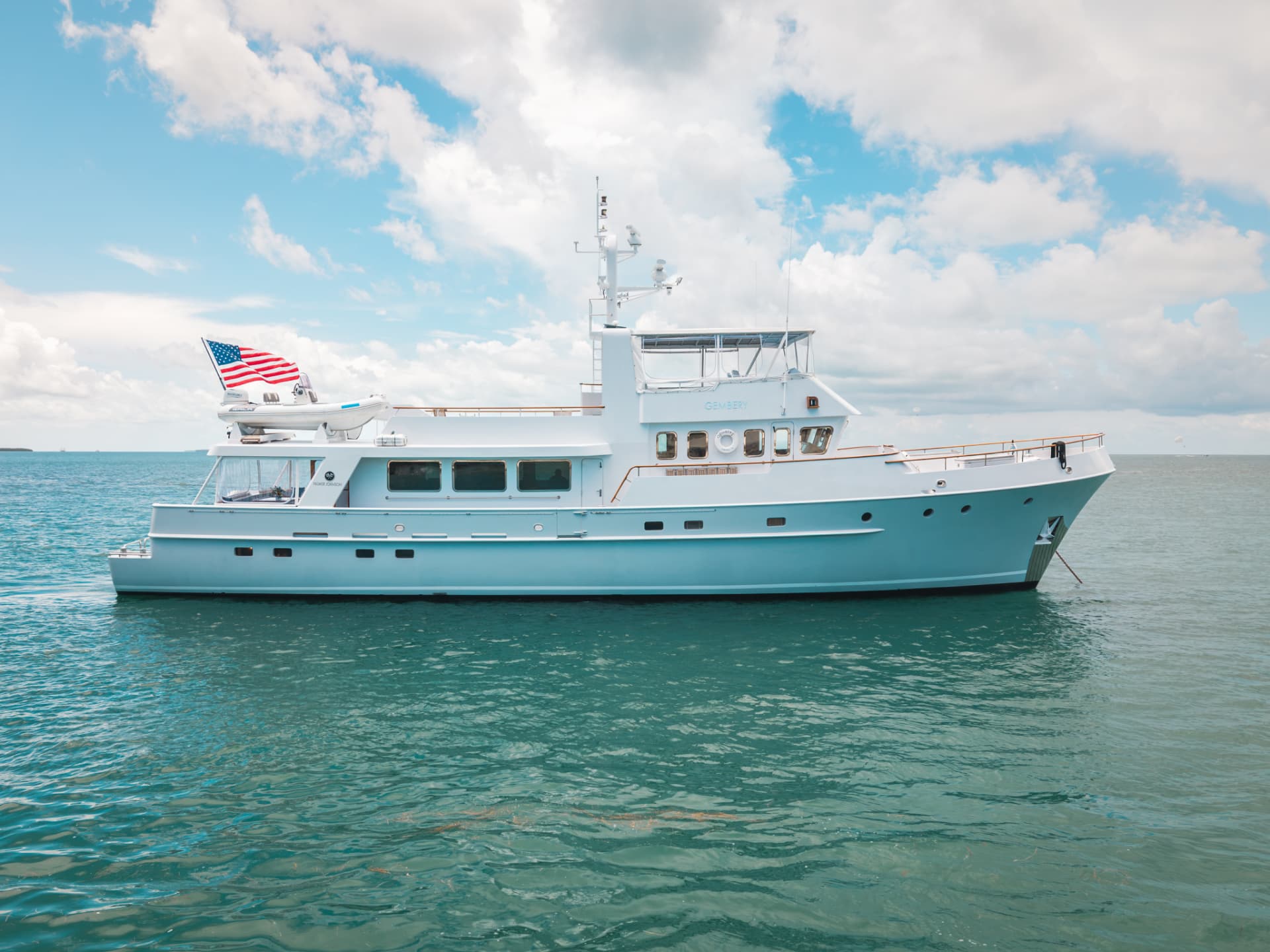 A light blue Palmer Johnson motor yacht at anchor in clear turquoise water under a partly cloudy sky
